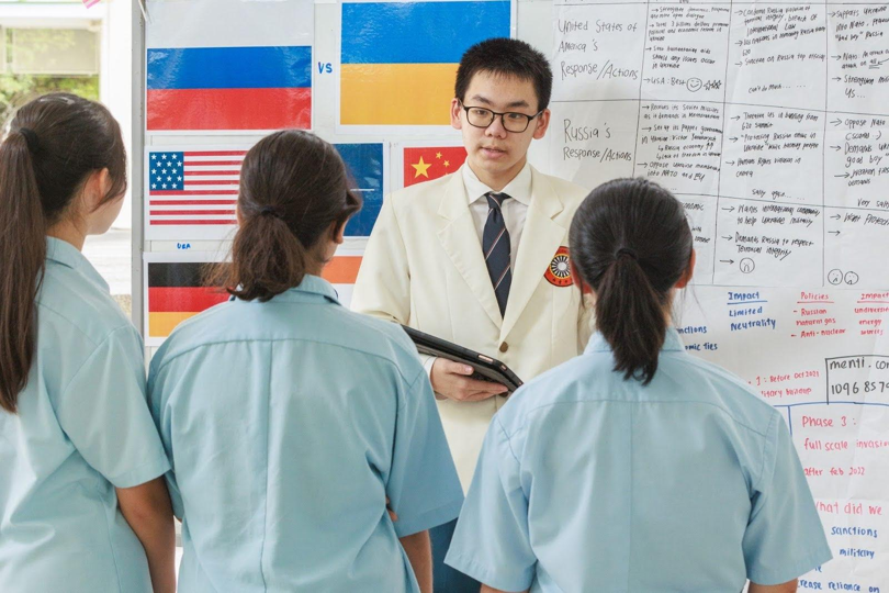 Student presents to classmates using a tablet in front of a classroom display with international flags and notes on global responses.